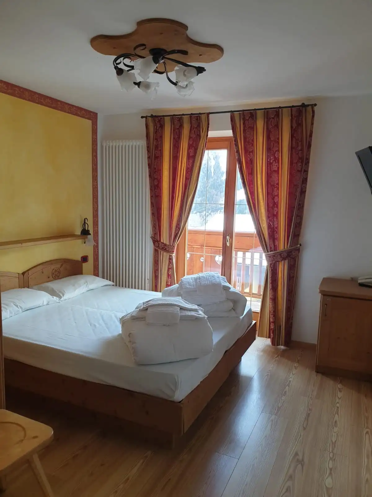 Letto matrimoniale con parete gialla, lampadario in legno e terrazzo affacciato sulla neve