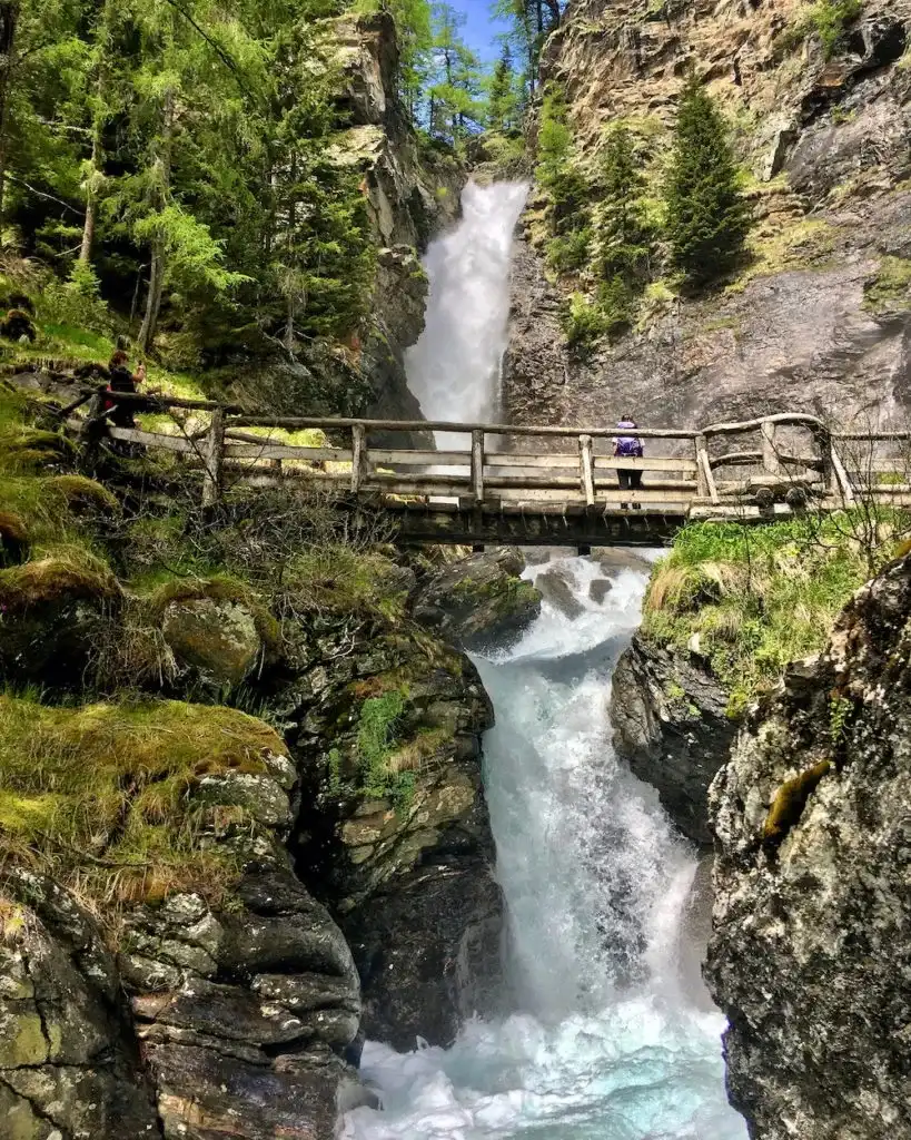 La cascata di Saènt che precipita tra le rocce, con il ponte di legno e il bosco