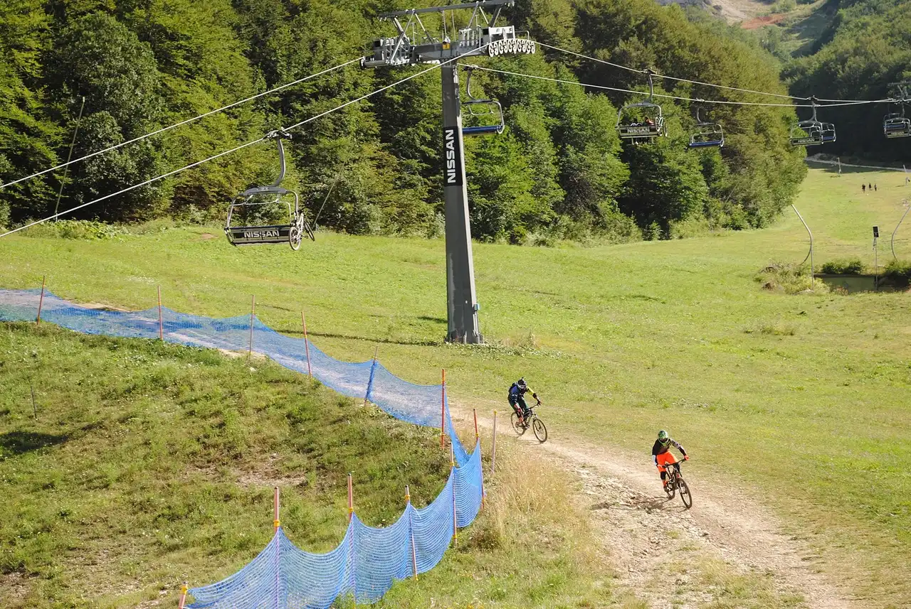 Due biker in discesa su un tracciato sterrato tra i prati, con la seggiovia che porta le bici a monte
